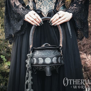 Puede incluir: Un bolso negro de cuero con forma de caldero, con asa y diseño decorativo de fases lunares. El bolso tiene un pico y lo sostiene una persona con un vestido negro. La marca "Otherworld Workshop" es visible.