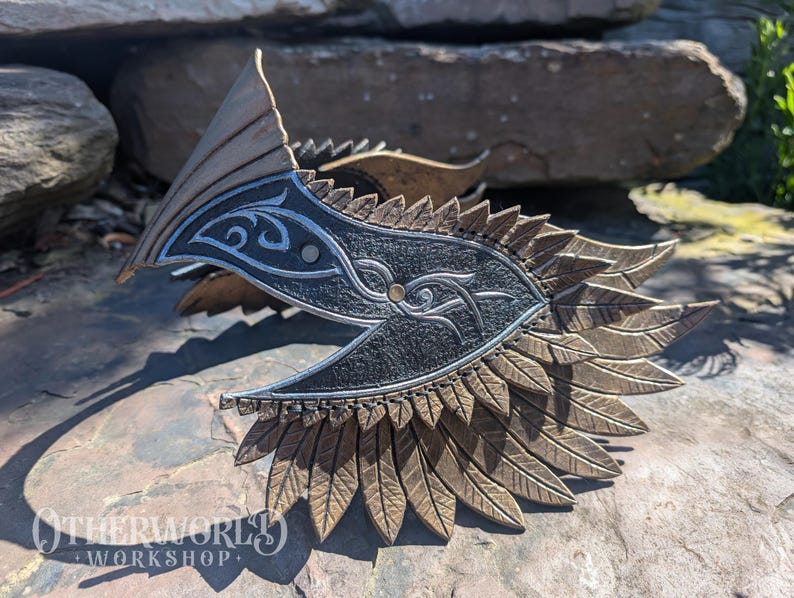 May include: A bronze-colored, decorative mask with a central black panel featuring silver scrollwork and an eye design. The mask has a feathered edge and a pointed top, resting on a stone surface. The logo "OTHERWORLD WORKSHOP" is visible.