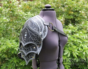 May include: Black and silver shoulder armor with intricate etched designs, including crescent moons and swirling patterns. The armor is attached to a black leather strap with a gold-colored ring. The shoulder armor is displayed on a mannequin.