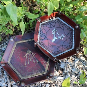 May include: Two hexagonal, leather-bound trays with decorative designs. One tray features a deer skull and crescent moon, the other a star and arrows. Both have a burgundy interior and brass-colored trim.