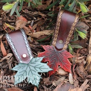 May include: Two leather keychains with leaf designs. One keychain features a red maple leaf, the other a green leaf. The keychains have brown leather straps with braided accents and a button closure. The background is composed of wood chips and foliage.