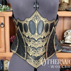 May include: Black and gold corset with intricate filigree and oval accents. The corset is displayed on a mannequin, showcasing its detailed design and craftsmanship. The corset has a diamond pattern on the lower section.