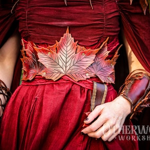 Puede incluir: Vestido burdeos con cinturón y brazaletes en forma de hojas de arce, hechos de cuero marrón y rojo. El fondo verde borroso resalta los detalles del vestido y los accesorios.