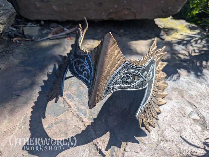 May include: A detailed, ornate headpiece with a metallic gold and silver finish. The design features intricate patterns and a central, pointed element. The headpiece is likely for costume or decorative purposes, with a shadow cast on the stone surface.