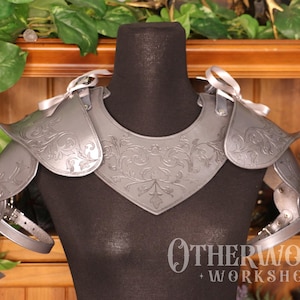 May include: Silver-colored shoulder armor with intricate floral designs, displayed on a black mannequin. The armor features multiple articulated plates and is secured with straps and ribbons. The product is from Otherworld Workshop.