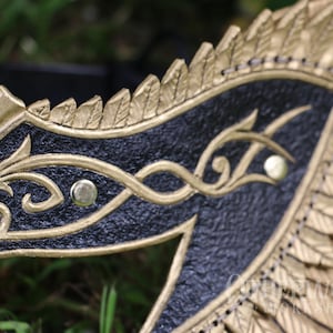 May include: Close-up of a gold and black mask with intricate detailing. The mask features a textured black surface with gold filigree designs and gold accents. The mask has a wing-like shape with feather details.
