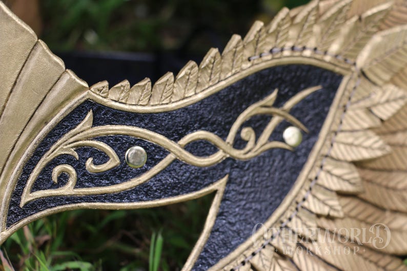 May include: Close-up of a gold and black mask with intricate detailing. The mask features a textured black surface with gold filigree designs and gold accents. The mask has a wing-like shape with feather details.