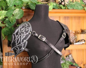 May include: Black leather shoulder armor with silver Celtic knotwork design. The armor is worn on a black mannequin and secured with adjustable straps and buckles. The image includes the text "OTHERWORLD WORKSHOP+".