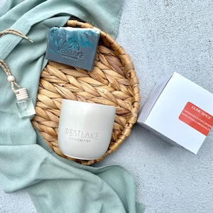 May include: A grey bar of soap with the Westlake Handmade logo, a white candle vessel with the Westlake Handmade logo, a small glass fragrance bottle with a wooden cap, and a white box labeled Dune Spice on a round woven tray with a sage green cloth.