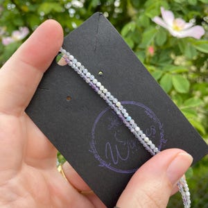 May include: Delicate beaded necklace with a mix of white, lavender, and pale blue beads. The necklace is displayed on a black card with a purple logo. The background features green foliage and a soft pink flower.
