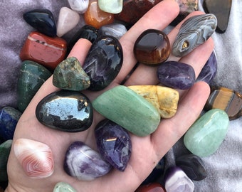 May include: A collection of polished stones in various colours and shapes, including green, purple, brown, red, and white. The stones are arranged in a hand, with some stones overlapping.
