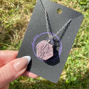 May include: A silver chain necklace featuring a raw, pink gemstone pendant. The gemstone is encased in silver wire and suspended from a delicate chain. The necklace is presented on a black card with a purple design, set against green grass.