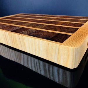 Op de afbeelding: Een rechthoekige houten snijplank met een lichtgekleurd frame en een donker, gestreept midden. Het midden heeft afwisselend donkere en lichte houtnerven. De plank heeft een gladde, gepolijste afwerking en een handgreep.