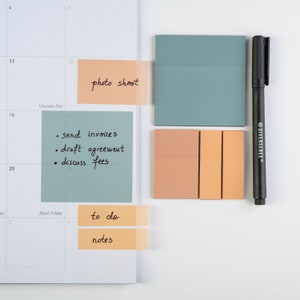 May include: A white calendar with sticky notes on it. The notes are in different colors and have handwritten text on them. The text on the notes includes "photo shoot", "send invoices", "draft agreement", "discuss fees", "to do", and "notes".