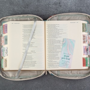 May include: A beige Bible cover with a zipper closure and two handles. The cover has a colorful geometric pattern. The Bible is open to the Psalms and Proverbs sections. A bookmark with the text "Above all else, guard your heart, for everything you do flows from it." is inserted in the Bible.
