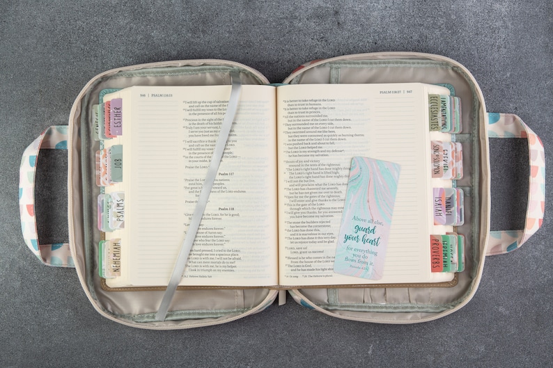May include: A beige Bible cover with a zipper closure and two handles. The cover has a colorful geometric pattern. The Bible is open to the Psalms and Proverbs sections. A bookmark with the text "Above all else, guard your heart, for everything you do flows from it." is inserted in the Bible.
