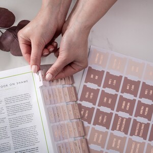 May include: A set of pink and beige bible tabs with gold lettering. The tabs are labeled with the names of the books of the Bible, including Genesis, Leviticus, Numbers, Deuteronomy, Joshua, Judges, Ruth, 1 Samuel, 2 Samuel, 1 Kings, 2 Kings, 1 Chronicles, 2 Chronicles, Ezra, Nehemiah, Esther, Job, Psalms, Proverbs, Ecclesiastes, Song of Solomon, Isaiah, Jeremiah, Lamentations, Ezekiel, Daniel, Hosea, Joel, Amos, Obadiah, Jonah, Micah, Nahum, Habakkuk, Zephaniah, Haggai, Zechariah, Malachi, Matthew, Mark, Luke, John, Acts, Romans, 1 Corinthians, 2 Corinthians, Galatians, Ephesians, Philippians, Colossians, 1 Thessalonians, 2 Thessalonians, 1 Timothy, 2 Timothy, Titus, Philemon, Hebrews, James, 1 Peter, 2 Peter, 1 John, 2 John, 3 John, Jude, Revelation.
