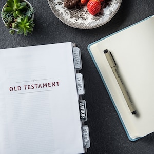 May include: A white book with the words "OLD TESTAMENT" printed on the cover. The book has tabs with the names of the first four books of the Old Testament: Genesis, Exodus, Leviticus, and Numbers.