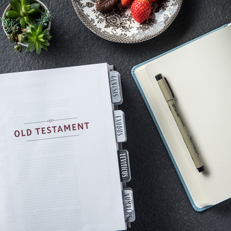 May include: A white book with the words "OLD TESTAMENT" printed on the cover. The book has tabs with the names of the first four books of the Old Testament: Genesis, Exodus, Leviticus, and Numbers.
