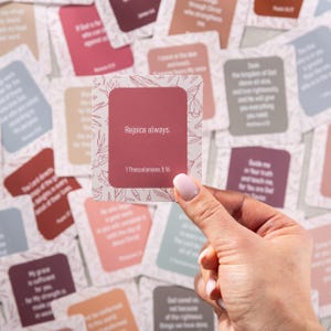 Pu&ograve; includere: Una mano che tiene in mano una carta quadrata con il testo "Rejoice always." e "1 Thessalonians 5:16" in un rettangolo rosso. La carta ha un design floreale sui bordi. Altre carte con testo sono sullo sfondo.