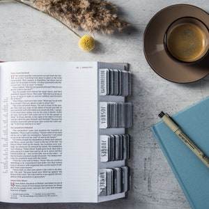 May include: A white open Bible with a black and white index tab system. The index tabs are labeled with the names of books of the Bible: Deuteronomy, Joshua, Judges, and Numbers. The Bible is open to Exodus 17, and the text is visible on the page.