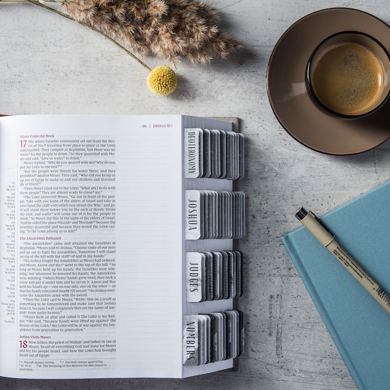 May include: A white open Bible with a black and white index tab system. The index tabs are labeled with the names of books of the Bible: Deuteronomy, Joshua, Judges, and Numbers. The Bible is open to Exodus 17, and the text is visible on the page.