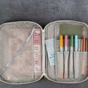 May include: An open beige zippered case with a green lining. The case has two handles and is filled with various items, including pens, pencils, and a small card with the text "Allow grace to guide your life".