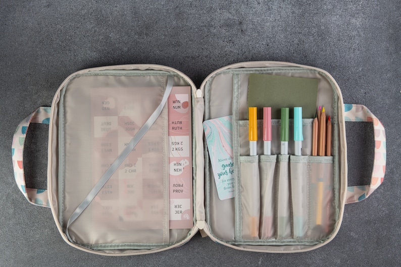 May include: An open beige zippered case with a green lining. The case has two handles and is filled with various items, including pens, pencils, and a small card with the text "Allow grace to guide your life".