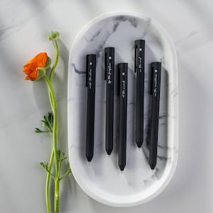 May include: Five black pens with white lettering on a marble surface. The pens have the following text: "into the storm", "by the fireflies", "at the sound", "on the wind", and "in the meantime".