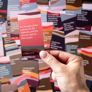 May include: A rectangular card with the text "For the eyes of the Lord are on the righteous, and His ears are open to their prayer. 1 Peter 3:12" is held in a hand. The card has a layered design with shades of red, pink, and brown.