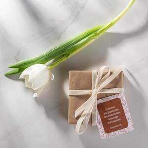 Pu&ograve; includere: Un regalo marrone avvolto, legato con un nastro color crema, accompagnato da un tulipano bianco e un'etichetta con testo. L'etichetta recita: "All'uomo che gli piace, Dio d&agrave; saggezza, conoscenza e felicit&agrave;. Ecclesiaste 2:26."