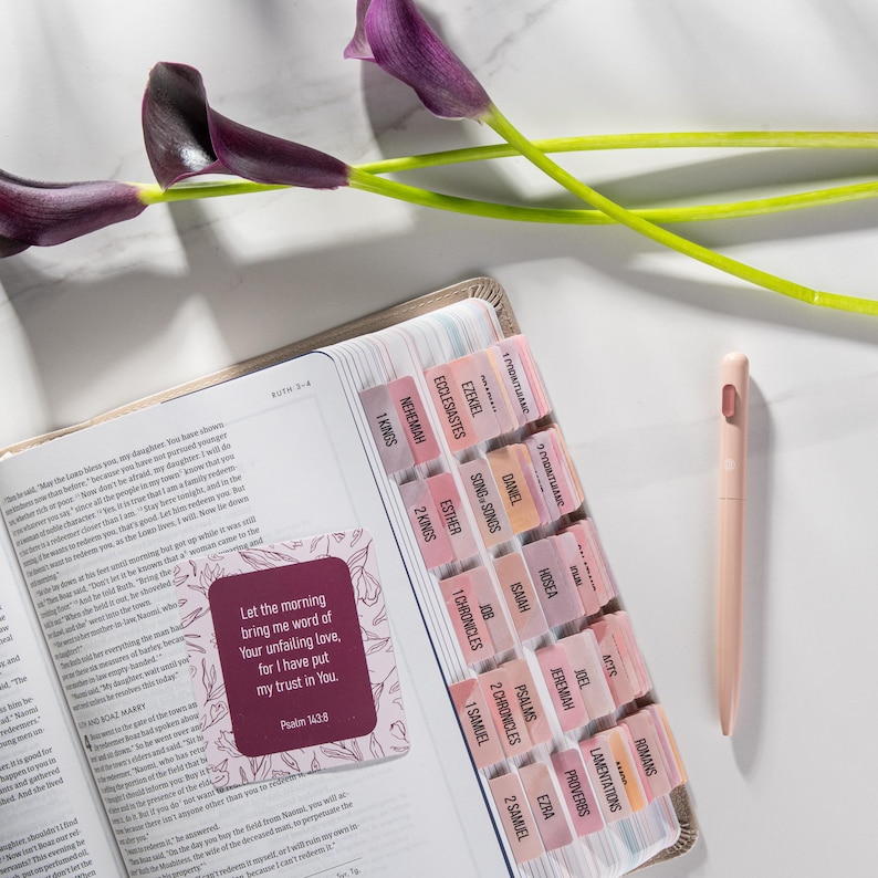 Pu&ograve; includere: Bibbia aperta con linguette rosa e pesca, una penna abbinata e una scheda decorativa con testo. Le linguette sono etichettate con i nomi dei libri. Le calle viola sono sullo sfondo.