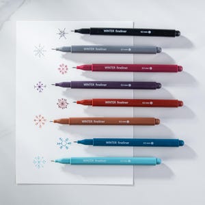 May include: A collection of seven Winter fineliner pens in various colors, including black, gray, burgundy, purple, red, brown, blue, and teal. Each pen has a 0.5 mm tip and is displayed with snowflake drawings.