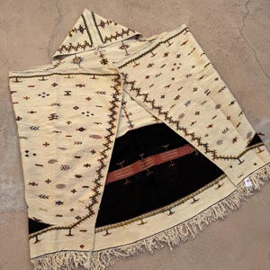 May include: Cream-colored hooded poncho with intricate geometric patterns in brown and red. The poncho features a black panel with a red stripe and fringe along the bottom edge. The design is a traditional textile.