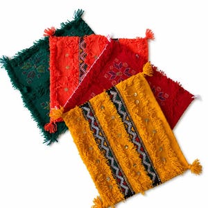 May include: A collection of four square textile mats in dark green, orange, red, and yellow. Each mat features vertical woven patterns, fringe edges, corner tassels, and small metallic circular embellishments sewn onto the fabric surface.