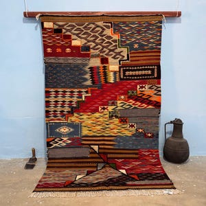 Op de afbeelding: Een rechthoekig geweven kleed met een complex patchwork van geometrische patronen in tinten rood, blauw, geel, bruin en zwart. Het kleed toont diverse getextureerde secties met zigzagpatronen, ruiten en strepen, afgewerkt met franjes aan de onderkant.