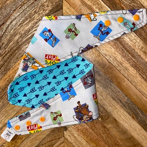 Op de afbeelding: Omkeerbare bandana met een lichtgrijze stof met tekenfilmfiguren en de tekst "TOYS PLAY". De achterkant is lichtblauw met een patroon van donkerblauwe pijlen. Met oranje drukknoopsluitingen.