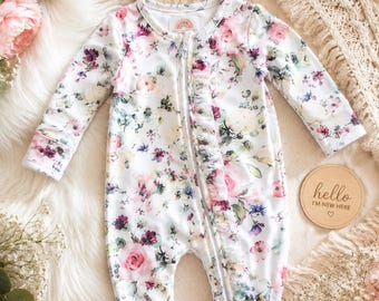 May include: A long-sleeved, footed infant sleeper with a white base and a floral pattern featuring pink, burgundy, and green flowers. The garment has a front zipper closure with a ruffled trim and a matching headband with a bow.