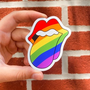 May include: A sticker featuring a stylized, rainbow-colored tongue and lips design. The design has a white border and is held against a red brick background. The colors of the rainbow are red, orange, yellow, green, blue, and purple.