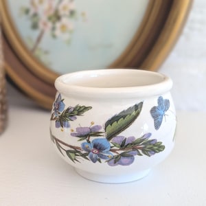 May include: A small, white ceramic pot with a floral and butterfly design. The pot features blue and purple flowers and a blue butterfly. The rim and base are plain white. The pot is likely for decorative use.