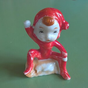 May include: A small ceramic figurine of a seated elf wearing a bright red suit and matching pointed hat. The elf has a white face with painted black eyes and a brown hair patch. It is posed with one arm raised and sits upon a textured base.