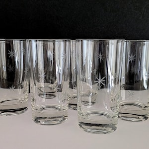 May include: Set of six clear, tall drinking glasses. Each glass features an etched starburst pattern. The glasses are cylindrical with a slightly flared base. They are displayed on a white surface against a dark background.