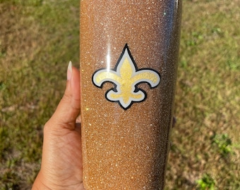 May include: A gold glitter tumbler with a black, white, and gold fleur-de-lis logo. The cylindrical tumbler has a clear lid. The background is blurred green grass.