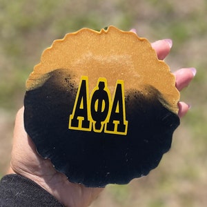 May include: A round coaster with a gold and black design. The top half is gold, and the bottom half is black. The letters "AΦA" are in gold on the black portion. The coaster has a wavy edge.