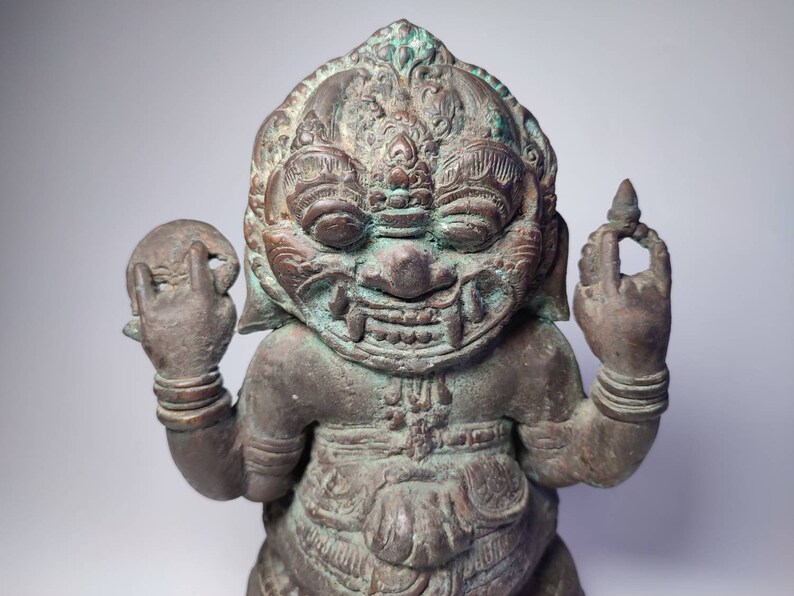 May include: A green and brown bronze statue of a mythical creature with a fierce expression. The statue has intricate details and is holding a round object in one hand and a small object in the other.