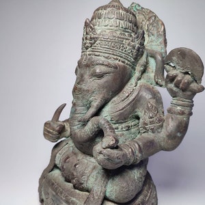 May include: A detailed bronze statue of Ganesha, the Hindu god of wisdom and prosperity, with a green patina. The statue depicts Ganesha seated with his trunk raised and holding a broken disc in his upper right hand.