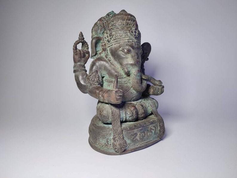 May include: A green bronze statue of Ganesha, the Hindu god of wisdom and prosperity, seated on a base with intricate carvings. The statue has a detailed headpiece and four arms, holding various objects.