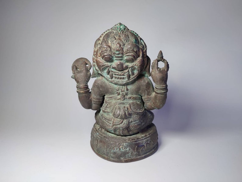 May include: A green bronze statue of a seated deity with a fierce expression. The deity has a crown and is holding a round object in one hand and a pointed object in the other.