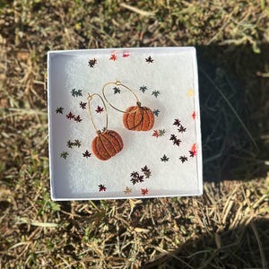May include: A pair of gold hoop earrings featuring orange glitter pumpkin charms, presented in a white box filled with small, scattered metallic maple leaf confetti. The pumpkin charms have a textured, sparkling finish and are attached to thin gold hoops.
