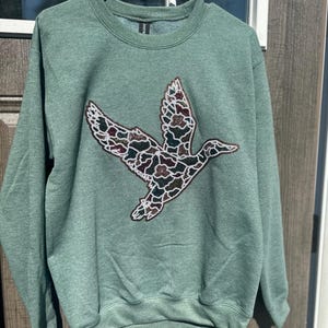 Op de afbeelding: Een saliegroene sweater met ronde hals en een grote, centrale applicatie van een vliegende eend. Het eendontwerp is gevuld met een meerkleurig camouflagepatroon bestaande uit kleine, glinsterende lovertjes in bruin, groen en wit.